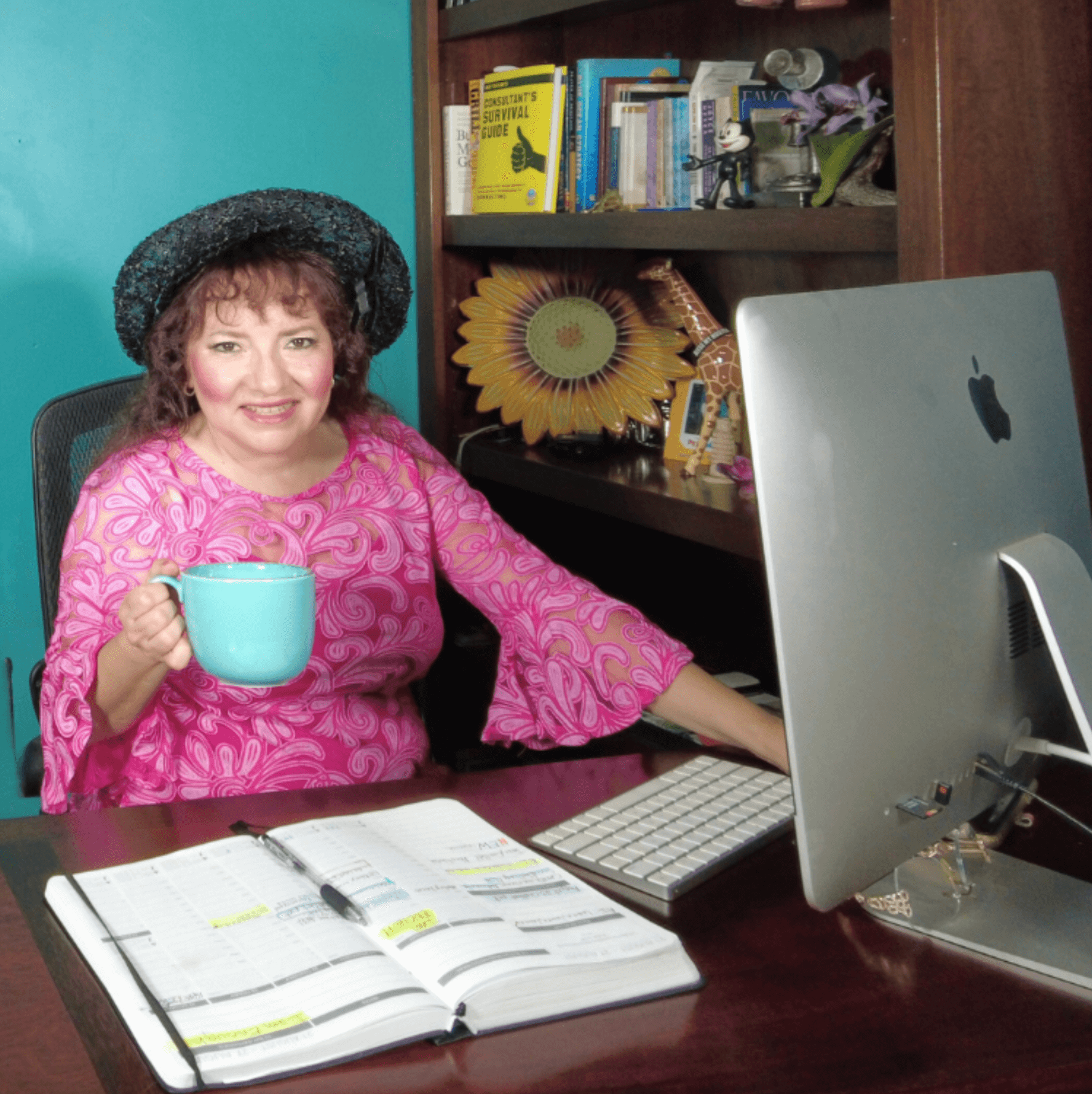 Gracie Ruth, LinkedIn Branding Expert and Personal Branding Coach, working at her desk with planner, computer, and coffee mug, in her teal-colored office with a sunflower on the wall